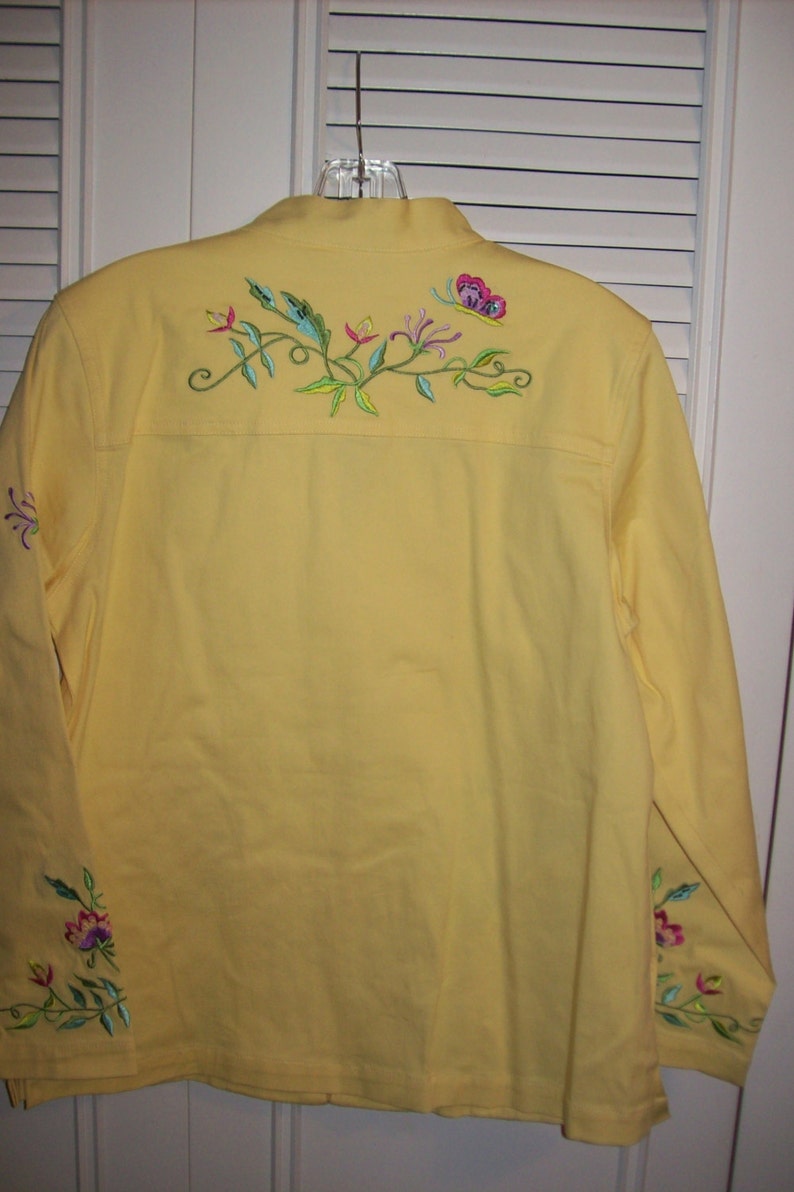 May include: A yellow jacket with long sleeves and a collar. The back of the jacket features embroidered floral and butterfly designs in pink, green, and blue. Additional floral embroidery is on the sleeves.