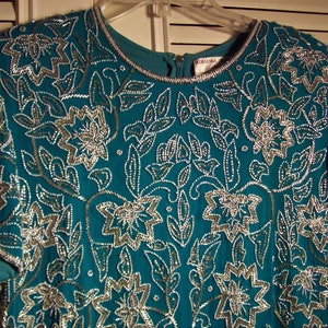 May include: Teal blue sequined top with a silver floral pattern. The top has a round neckline and short sleeves.
