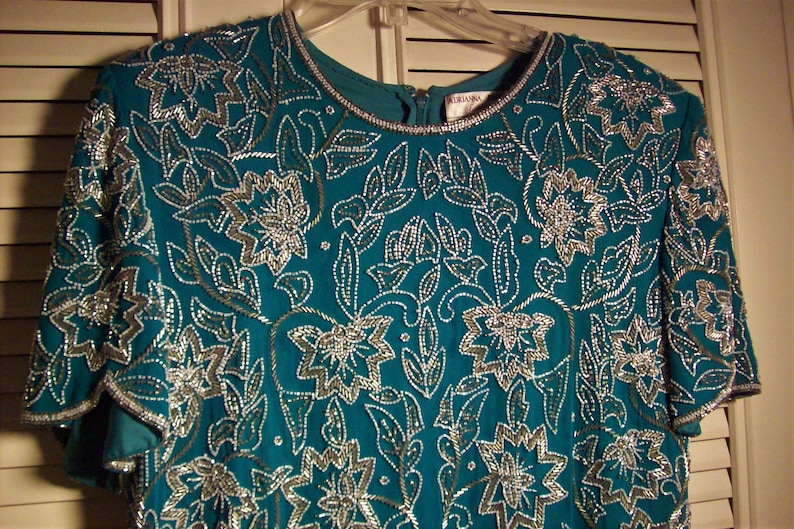 May include: Teal blue sequined top with a silver floral pattern. The top has a round neckline and short sleeves.