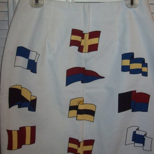 May include: White skirt adorned with colorful nautical flag designs. The flags feature various color combinations, including red, yellow, blue, and black. The skirt is hanging on a clear plastic hanger.