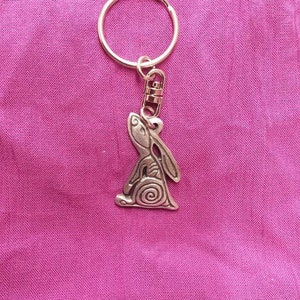 Puede incluir: Un llavero de plata con un dije de conejo detallado. El conejo está mirando hacia arriba con las orejas levantadas. El llavero está sobre un fondo de tela rosa.