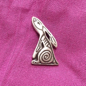 May include: A silver-coloured metal rabbit-shaped pin. The pin has a stylised design with a spiral at the base and a detailed outline. The pin is set against a textured, magenta-coloured fabric background.