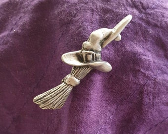 Puede incluir: Una figura de bruja plateada con un sombrero puntiagudo y una escoba. La figura está volando por el aire.