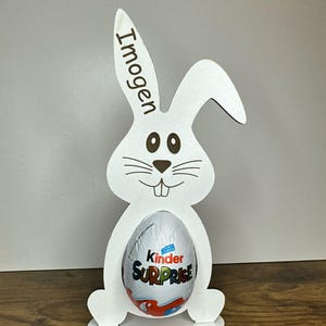 K&ouml;nnte beinhalten: Wei&szlig;er h&ouml;lzerner Osterhasen-Eierhalter mit dem Namen "Imogen" auf dem Ohr graviert. Der Hase h&auml;lt ein Kinder &Uuml;berraschungsei.