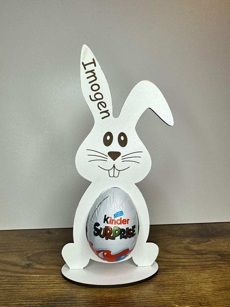K&ouml;nnte beinhalten: Wei&szlig;er h&ouml;lzerner Osterhasen-Eierhalter mit dem Namen "Imogen" auf dem Ohr graviert. Der Hase h&auml;lt ein Kinder &Uuml;berraschungsei.