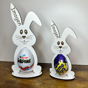 K&ouml;nnte beinhalten: Zwei wei&szlig;e h&ouml;lzerne Osterhasen-Eierhalter mit dem Namen "Imogen" auf jedem graviert. Der gr&ouml;&szlig;ere Hase h&auml;lt ein Kinder &Uuml;berraschungsei und der kleinere Hase h&auml;lt ein Cadbury Creme Ei.