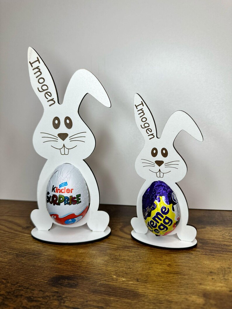 K&ouml;nnte beinhalten: Zwei wei&szlig;e h&ouml;lzerne Osterhasen-Eierhalter mit dem Namen "Imogen" auf jedem graviert. Der gr&ouml;&szlig;ere Hase h&auml;lt ein Kinder &Uuml;berraschungsei und der kleinere Hase h&auml;lt ein Cadbury Creme Ei.