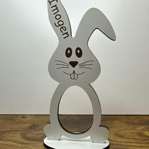 K&ouml;nnte beinhalten: Wei&szlig;er h&ouml;lzerner Osterhasen-Eierhalter mit dem Namen "Imogen" auf dem Ohr graviert. Der Hase hat braune Augen und eine braune Nase.