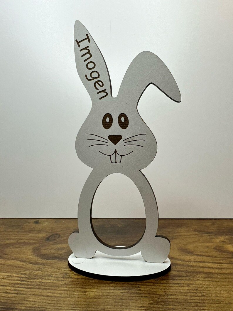 K&ouml;nnte beinhalten: Wei&szlig;er h&ouml;lzerner Osterhasen-Eierhalter mit dem Namen "Imogen" auf dem Ohr graviert. Der Hase hat braune Augen und eine braune Nase.