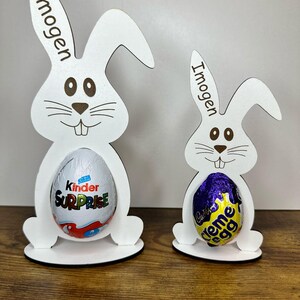 K&ouml;nnte beinhalten: Zwei wei&szlig;e Holz-Hasen-Eierhalter mit dem Namen Imogen auf jedem. Der gr&ouml;&szlig;ere Hase h&auml;lt ein Kinder &Uuml;berraschungsei und der kleinere Hase h&auml;lt ein Cadbury Creme Ei.
