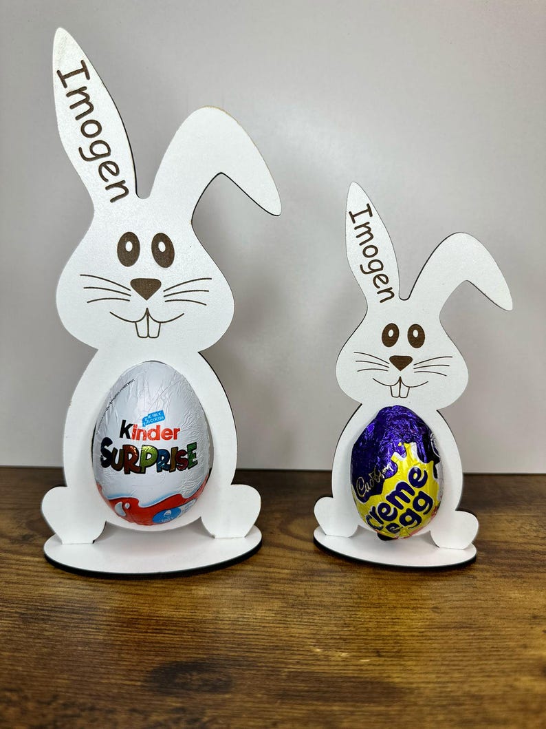 K&ouml;nnte beinhalten: Zwei wei&szlig;e Holz-Hasen-Eierhalter mit dem Namen Imogen auf jedem. Der gr&ouml;&szlig;ere Hase h&auml;lt ein Kinder &Uuml;berraschungsei und der kleinere Hase h&auml;lt ein Cadbury Creme Ei.