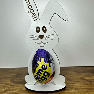 K&ouml;nnte beinhalten: Wei&szlig;er h&ouml;lzerner Osterhasen-Eierhalter mit dem Namen "Imogen" auf dem Ohr graviert. Der Hase h&auml;lt ein lila und gelbes, in Folie verpacktes Schokoladenei.
