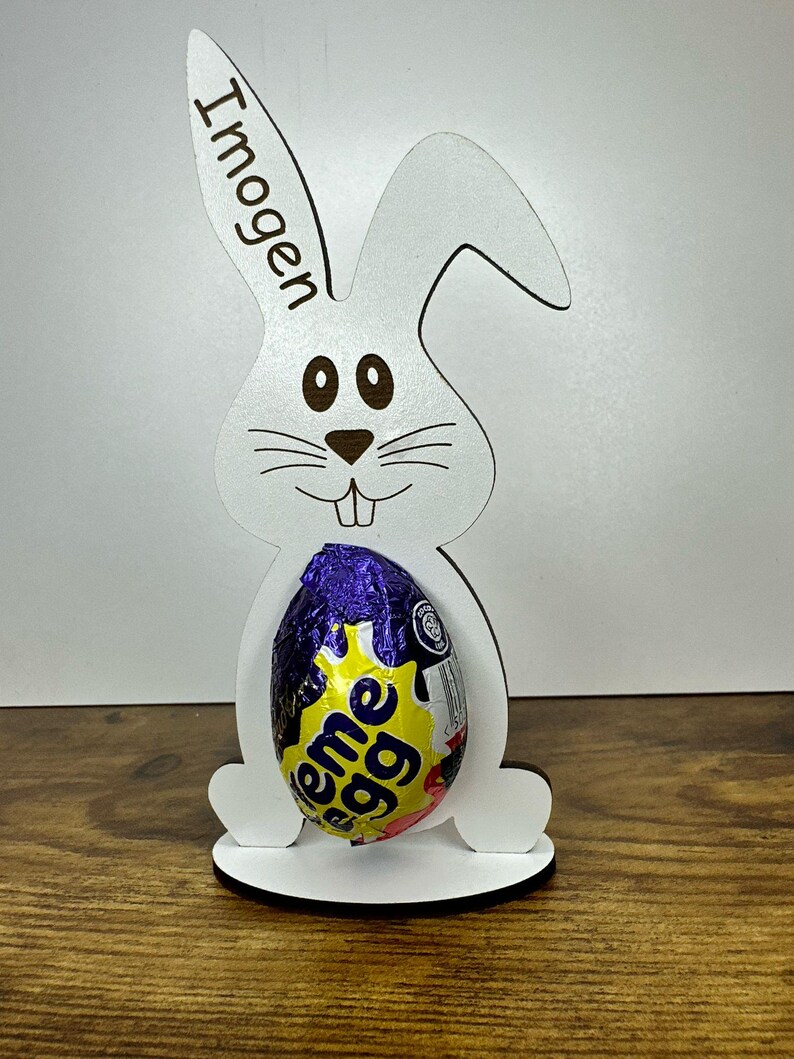 K&ouml;nnte beinhalten: Wei&szlig;er h&ouml;lzerner Osterhasen-Eierhalter mit dem Namen "Imogen" auf dem Ohr graviert. Der Hase h&auml;lt ein lila und gelbes, in Folie verpacktes Schokoladenei.