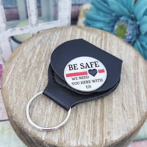 May include: Black leather keychain with a silver charm that reads "Be Safe We Need You Here With Us" with a red stripe and a black heart.