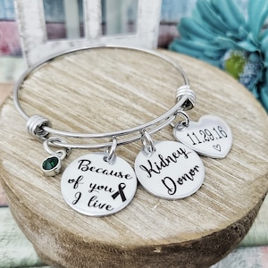 May include: Silver charm bracelet with three charms. The charms say "Because of you I live" with a black ribbon, "Kidney Donor" and "11.29.16" with a heart. The bracelet has a green crystal charm.