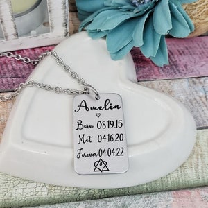 May include: Silver necklace with a rectangular tag that reads "Amelia" with a heart symbol, "Born 08.19.15", "Met 04.16.20", "Forever 04.04.22", and a symbol of three interlocking circles.