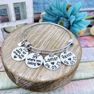 May include: A silver bangle bracelet with four charms. The charms have the text "It's not where you come from", "It's where you belong", "Lamar 06.03.22", and "Revon 06.03.22".