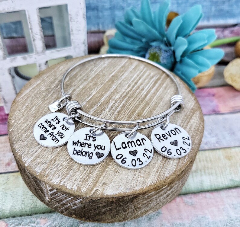 May include: A silver bangle bracelet with four charms. The charms have the text "It's not where you come from", "It's where you belong", "Lamar 06.03.22", and "Revon 06.03.22".