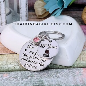 May include: Silver keychain with two round charms. The larger charm has the text "Remember the joy, enjoy the present, embrace the future". The smaller charm has the text "40 Years" and a small apple and book graphic. A pink rhinestone is attached to the top of the keychain.