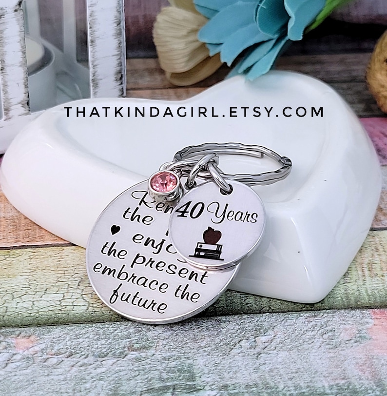 May include: Silver keychain with two round charms. The larger charm has the text "Remember the joy, enjoy the present, embrace the future". The smaller charm has the text "40 Years" and a small apple and book graphic. A pink rhinestone is attached to the top of the keychain.
