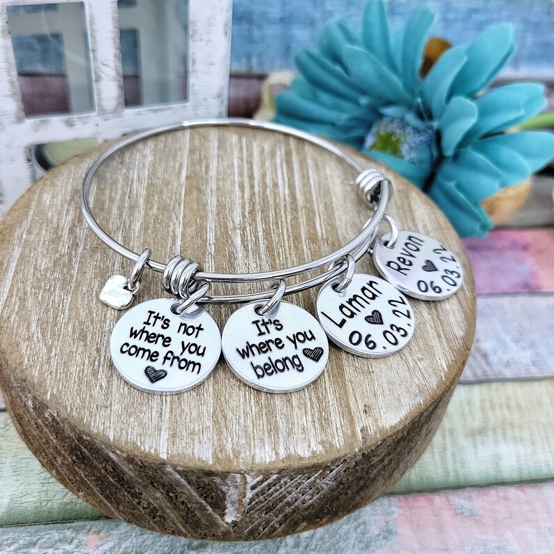 May include: A silver bangle bracelet with four charms. The charms have the text "It's not where you come from", "It's where you belong", "Lamar 06.03.22", and "Revon 06.03.22".