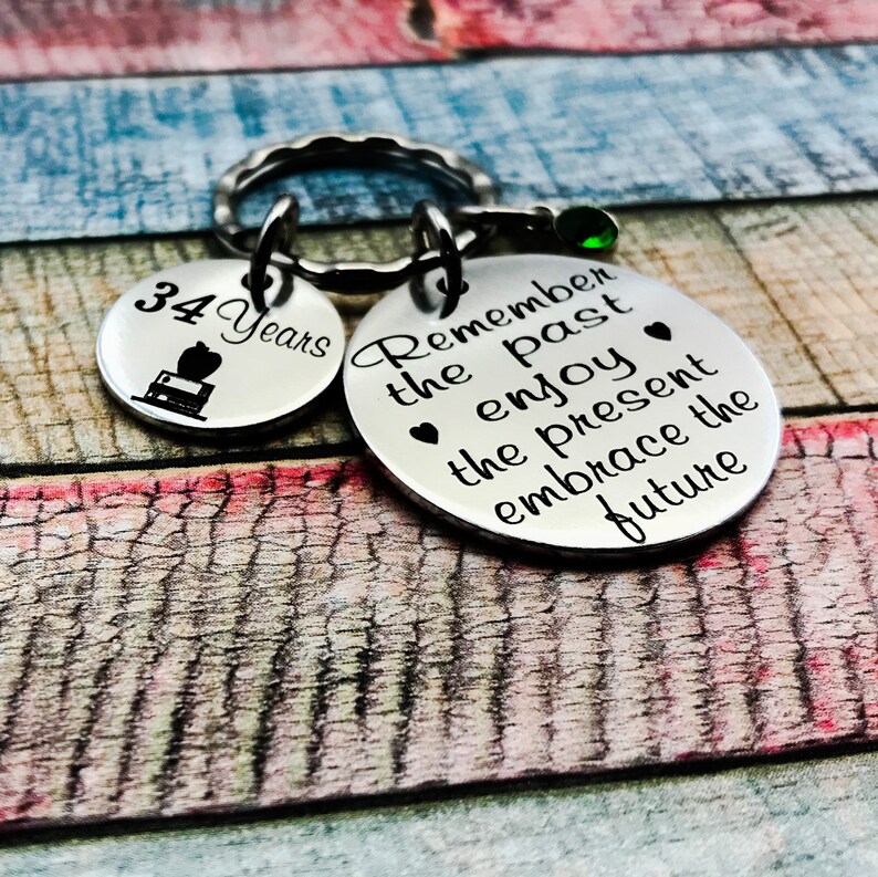 May include: A silver keychain with two round charms. The larger charm has the text "Remember the past enjoy the present embrace the future" with a small heart symbol before the words "enjoy" and "present". The smaller charm is silver with black text that reads "34 Years" and a small black image of an apple, a book, and a graduation cap.