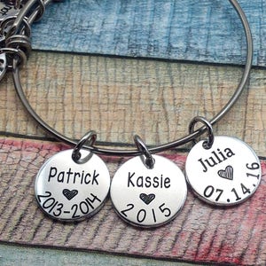 May include: A silver bangle bracelet with three charms. The charms are round and engraved with the names Patrick, Kassie, and Julia, along with dates and hearts.