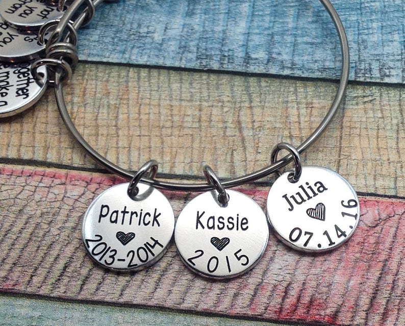 May include: A silver bangle bracelet with three charms. The charms are round and engraved with the names Patrick, Kassie, and Julia, along with dates and hearts.