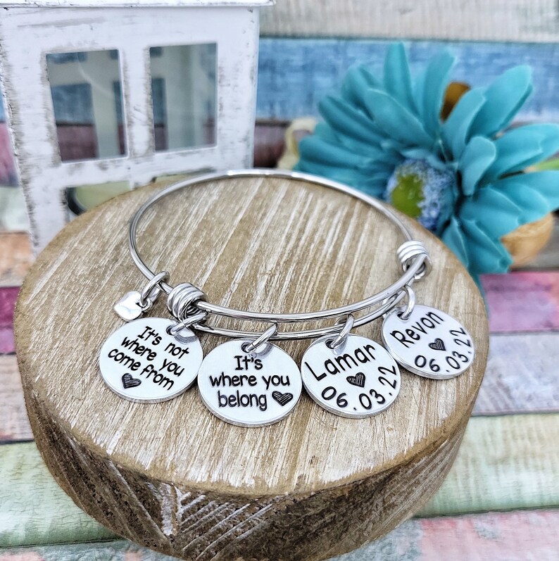 May include: Silver charm bracelet with four round charms. The charms have the text "It's not where you come from", "It's where you belong", "Lamar 06.03.22", and "Revon 06.03.22".