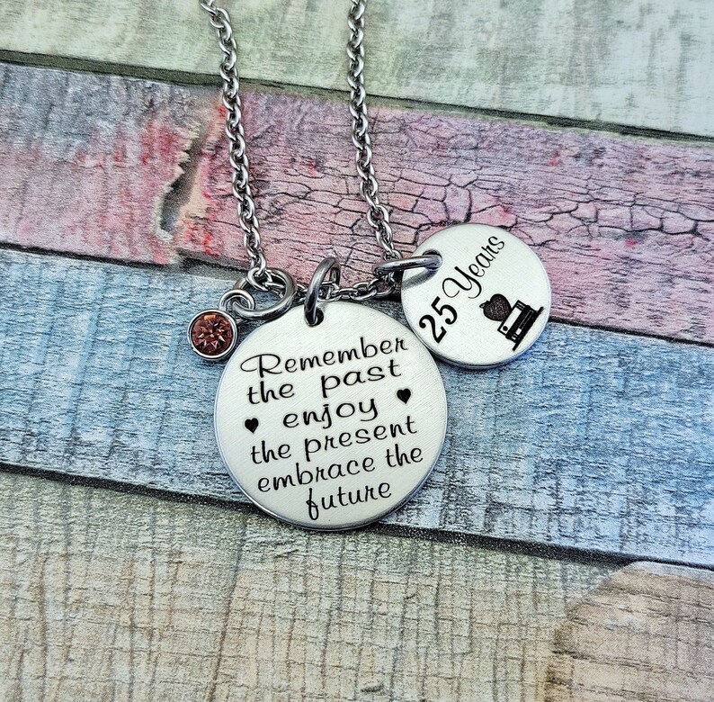 May include: Silver necklace with two round charms. The larger charm has the text "Remember the past enjoy the present embrace the future". The smaller charm has the text "25 Years" and a small image of a book.