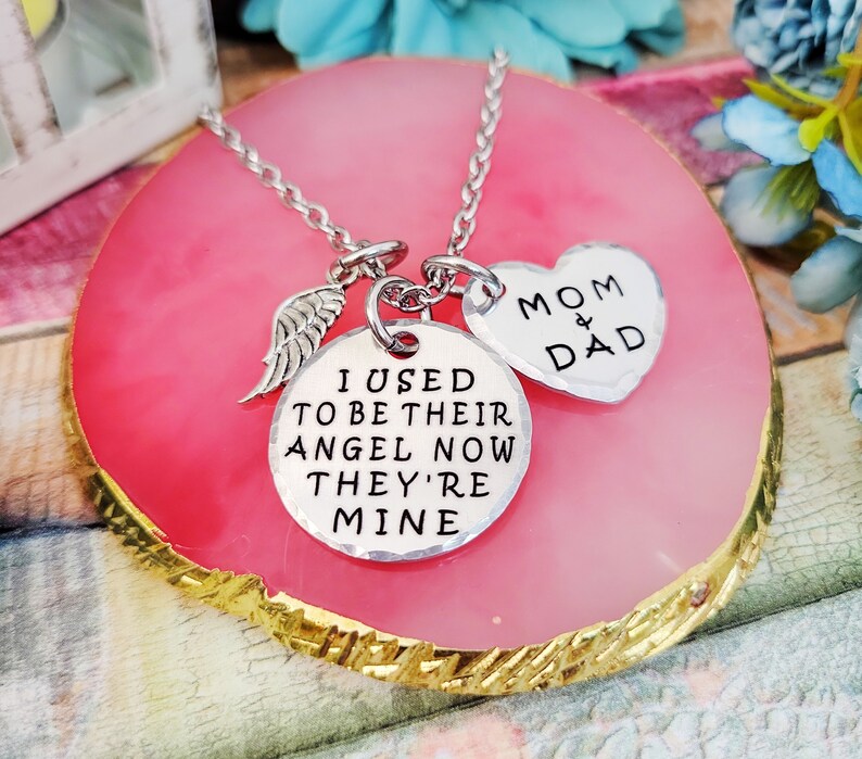 May include: Silver necklace with a heart charm that says "Mom & Dad" and a round charm that says "I used to be their angel now they're mine". The necklace also has a small angel wing charm.