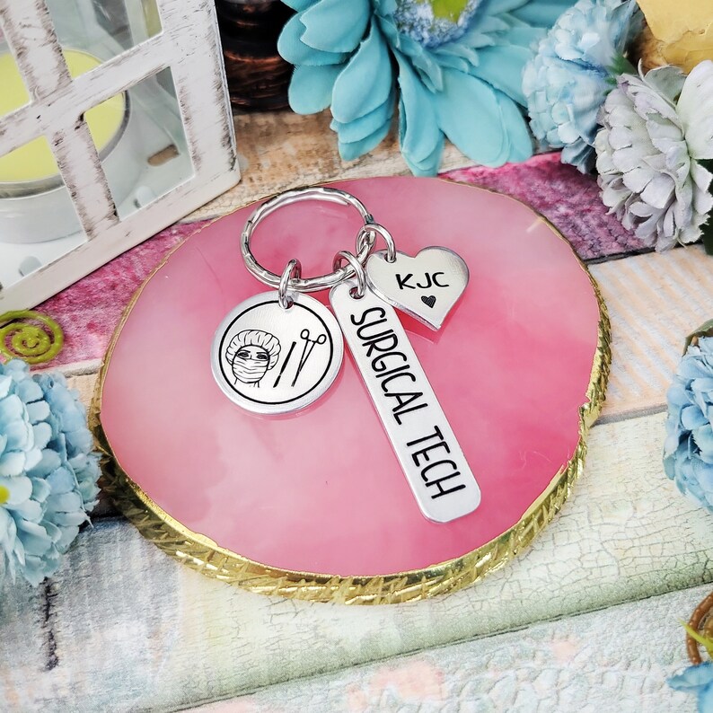 May include: Silver keychain with a surgical tech charm, a heart charm with the initials KJC, and a key ring. The charms are on a pink and gold background.