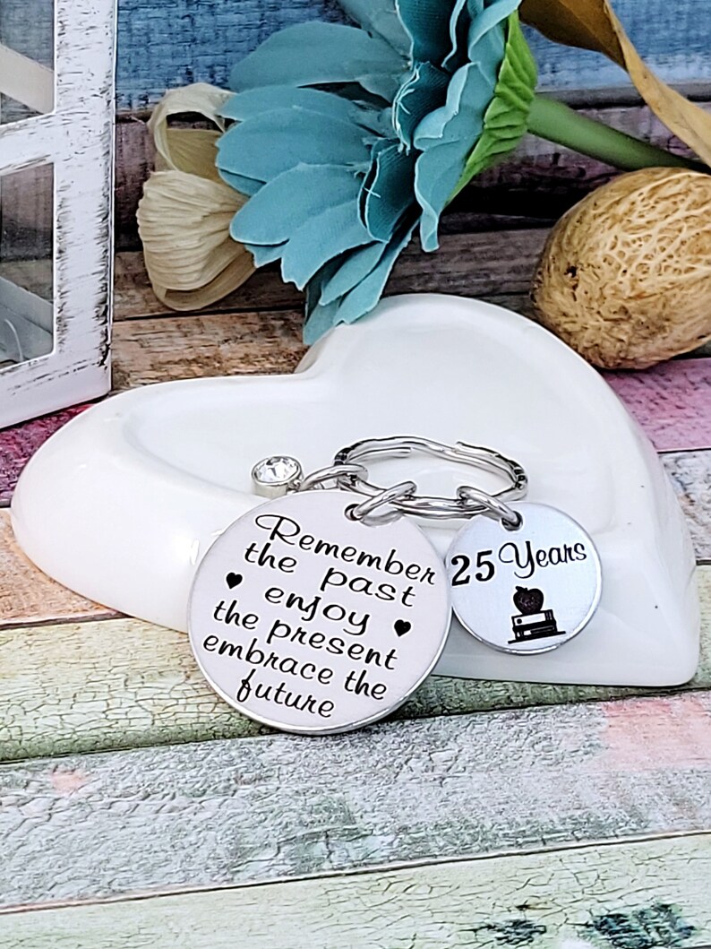 May include: Silver keychain with two round charms. One charm says "Remember the past enjoy the present embrace the future" and the other charm says "25 Years" with a book and apple graphic.