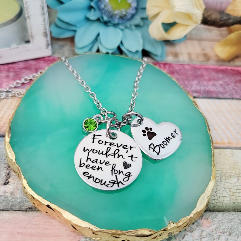 May include: Silver necklace with two charms. One charm is a round disc with the text "Forever wouldn't have been long enough" and a small heart. The other charm is a heart-shaped disc with a paw print and the name "Boomer". A green crystal is attached to the necklace chain.