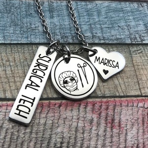 May include: A silver necklace with four charms. The charms are a rectangular tag that says "SURGICAL TECH", a round charm with a drawing of a person wearing a surgical cap and mask, a pair of surgical scissors, and a heart-shaped charm that says "MARISSA".