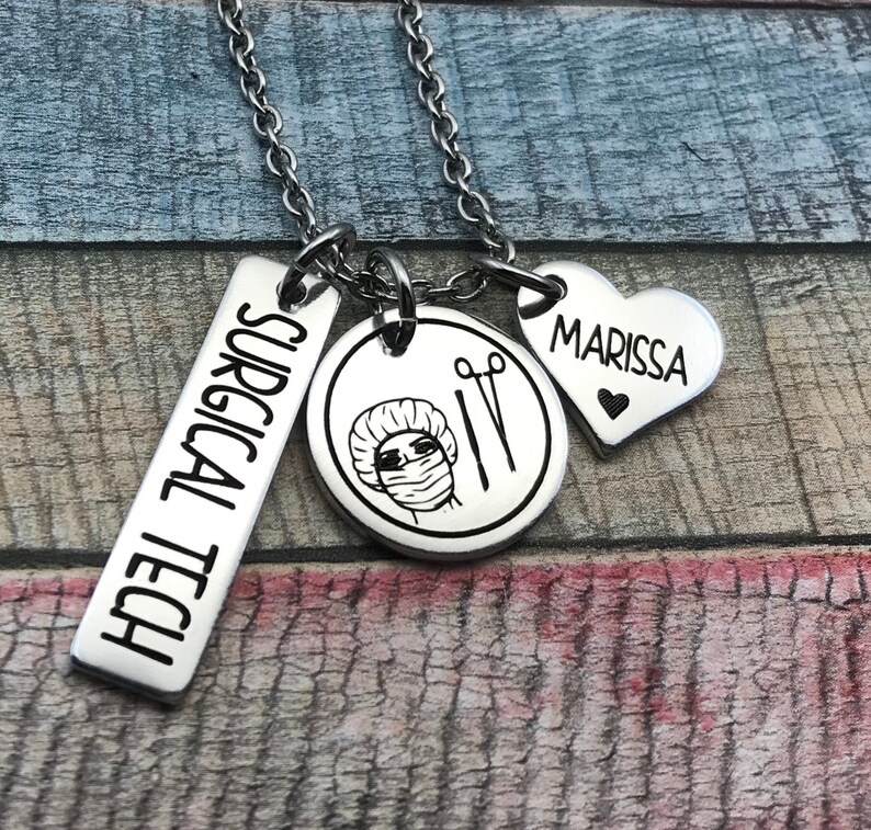 May include: A silver necklace with four charms. The charms are a rectangular tag that says "SURGICAL TECH", a round charm with a drawing of a person wearing a surgical cap and mask, a pair of surgical scissors, and a heart-shaped charm that says "MARISSA".