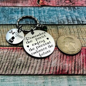May include: Silver keychain with a round charm that says "Remember the past enjoy the present embrace the future". There is a smaller charm that says "34 Years" with a small image of a cake.