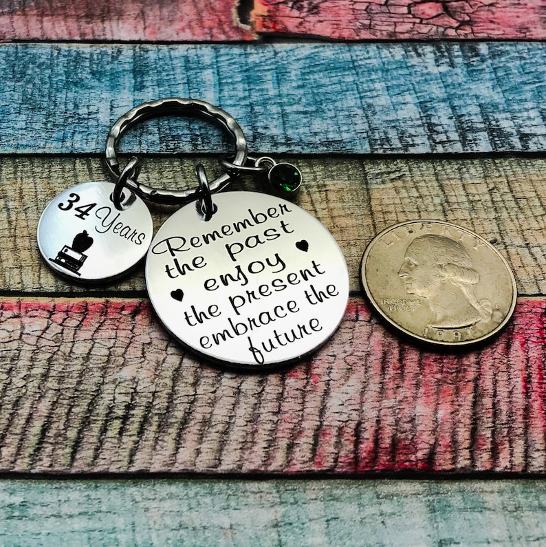 May include: Silver keychain with a round charm that says "Remember the past enjoy the present embrace the future". There is a smaller charm that says "34 Years" with a small image of a cake.