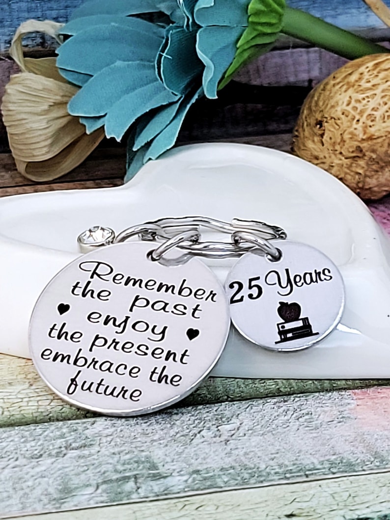 May include: Two silver keychains with a silver chain connecting them. One keychain has the text "Remember the past enjoy the present embrace the future" and the other keychain has the text "25 Years" with a small apple and books graphic.