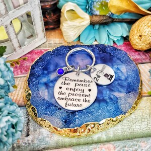 May include: A silver keychain with a blue gem charm and two round metal tags. The larger tag has the text "Remember the past enjoy the present embrace the future". The smaller tag has the text "24 Years" and a small graphic of a graduation cap. The keychain is on a blue and gold agate coaster.