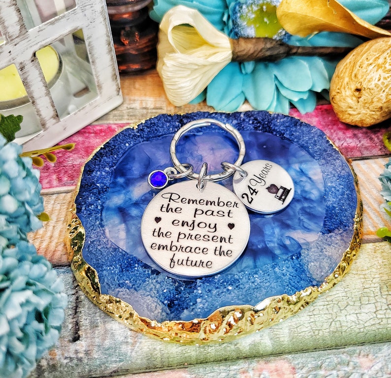 May include: A silver keychain with a blue gem charm and two round metal tags. The larger tag has the text "Remember the past enjoy the present embrace the future". The smaller tag has the text "24 Years" and a small graphic of a graduation cap. The keychain is on a blue and gold agate coaster.