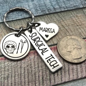 May include: Silver keyring with a surgical tech charm, a heart charm with the name Marissa, and a round charm featuring a surgical mask and tools. The keyring is attached to a silver key ring.