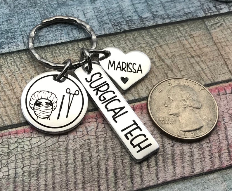 May include: Silver keyring with a surgical tech charm, a heart charm with the name Marissa, and a round charm featuring a surgical mask and tools. The keyring is attached to a silver key ring.