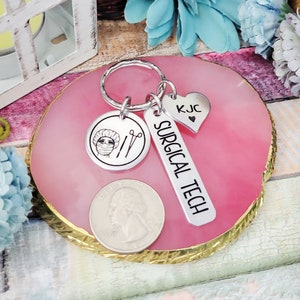 May include: Silver keyring with a surgical tech charm, a heart charm with the initials KJC, and a round charm featuring a surgical cap and scissors. The charms are attached to a silver key ring.
