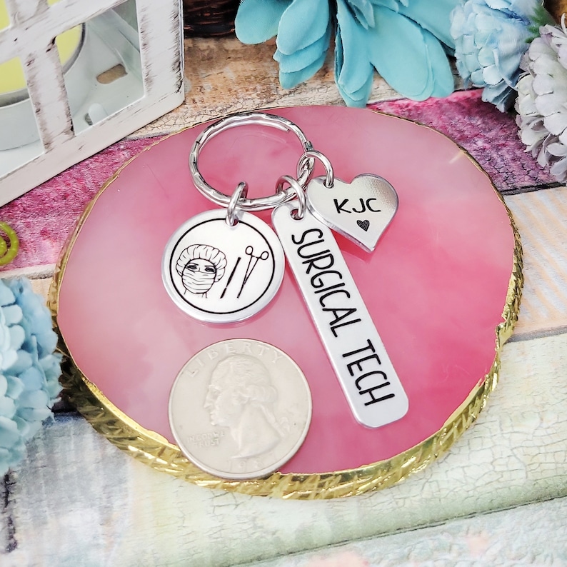May include: Silver keyring with a surgical tech charm, a heart charm with the initials KJC, and a round charm featuring a surgical cap and scissors. The charms are attached to a silver key ring.