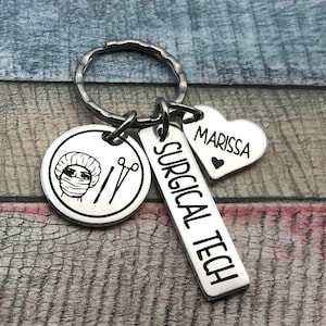 May include: Silver keyring with three charms: a round charm featuring a surgical technician wearing a surgical cap and mask, a rectangular charm with the text "SURGICAL TECH", and a heart-shaped charm with the text "MARISSA".