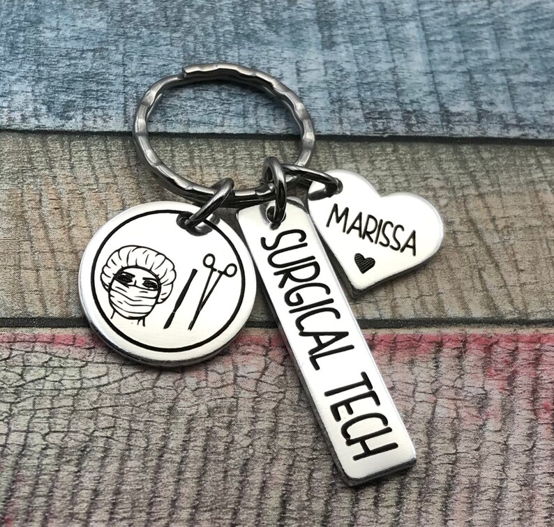 May include: Silver keyring with three charms: a round charm featuring a surgical technician wearing a surgical cap and mask, a rectangular charm with the text "SURGICAL TECH", and a heart-shaped charm with the text "MARISSA".