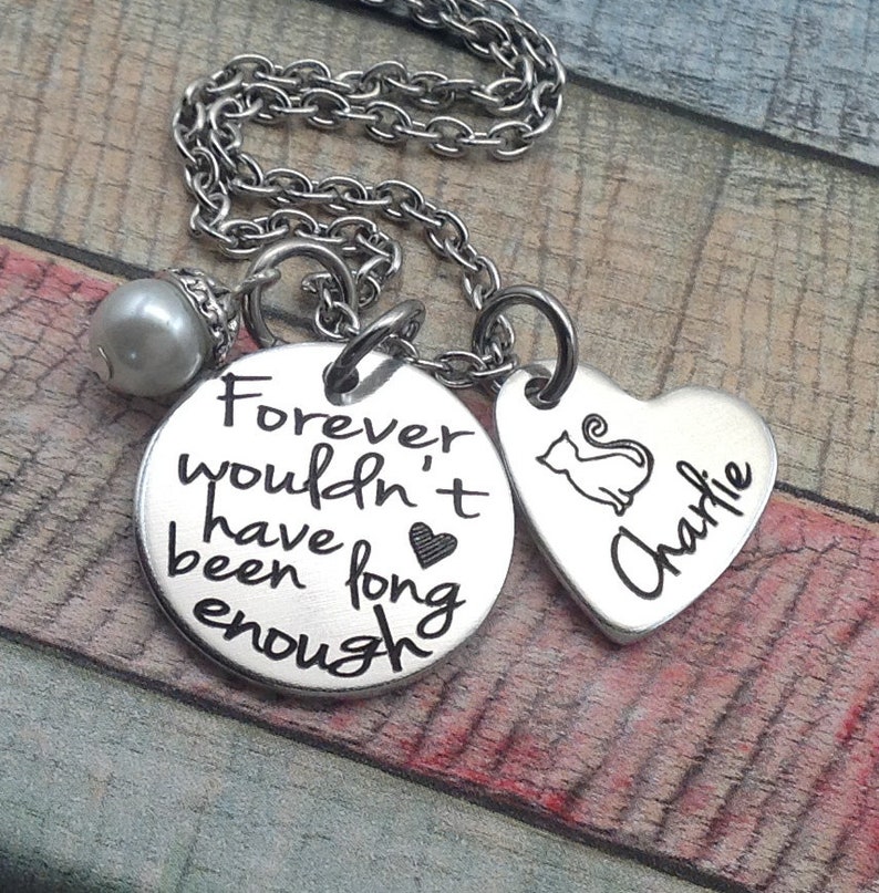 May include: Silver necklace with two charms. One charm is a round disc with the text "Forever wouldn't have been long enough" and a small heart. The other charm is a heart-shaped disc with a cat silhouette and the name "Charlie" engraved on it.