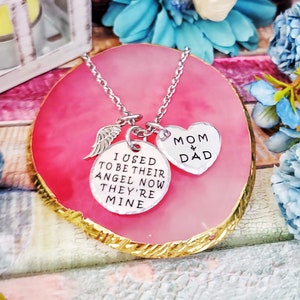 May include: Silver necklace with a heart charm that says "Mom & Dad" and a round charm that says "I used to be their angel now they're mine". The necklace is on a pink and gold background.