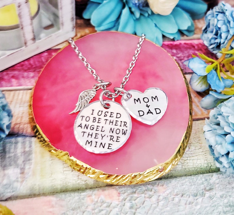 May include: Silver necklace with a heart charm that says "Mom & Dad" and a round charm that says "I used to be their angel now they're mine". The necklace is on a pink and gold background.
